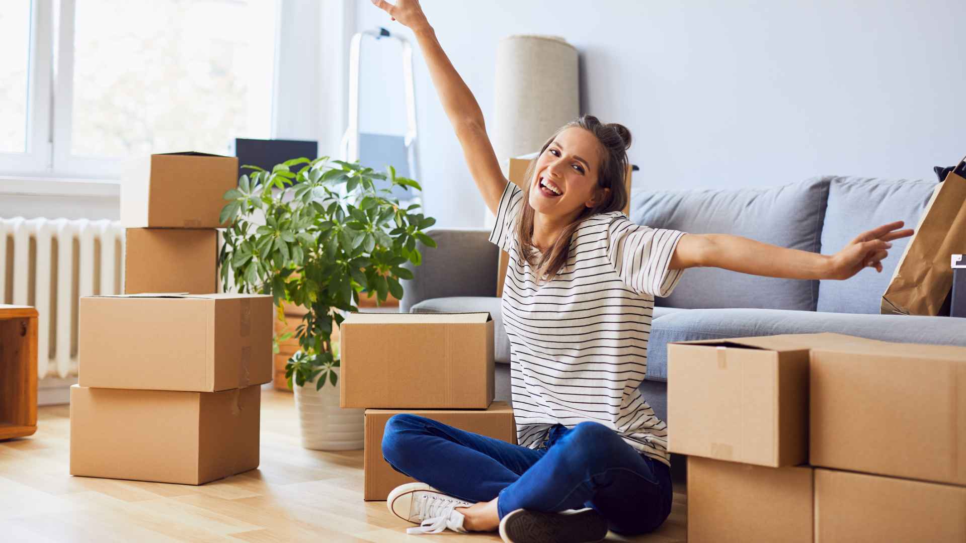 Young woman sitting in new apartment and raising arms in joy after moving in.